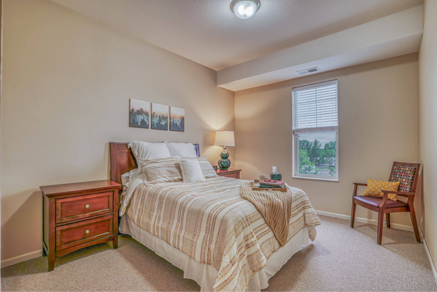 Cozy senior living unit with a neatly made bed, nightstands, and a chair by a window.