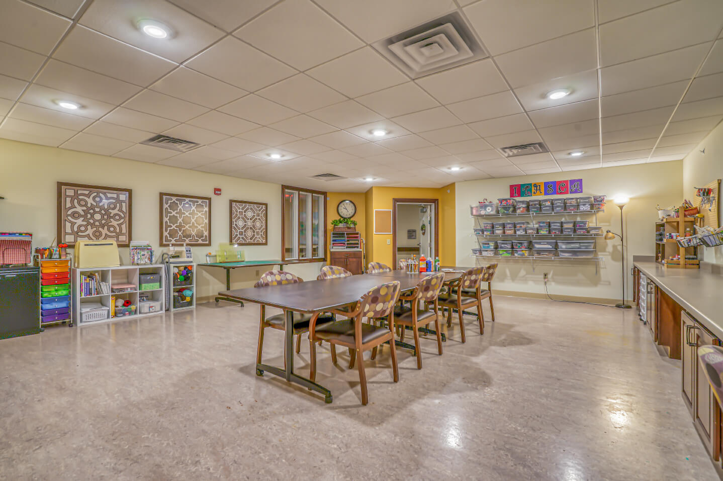 Well-lit activity room with tables, chairs, storage shelves, and art supplies.