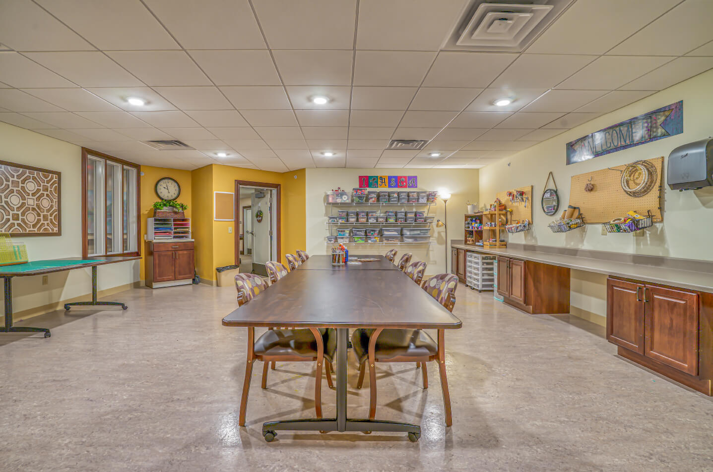 Well-lit craft room with tables, chairs, and organized shelves in a senior living unit.