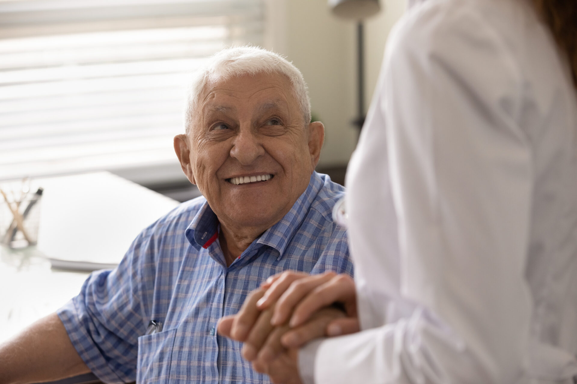 Elderly man smiling while holding hands with a caregiver in a senior living unit.