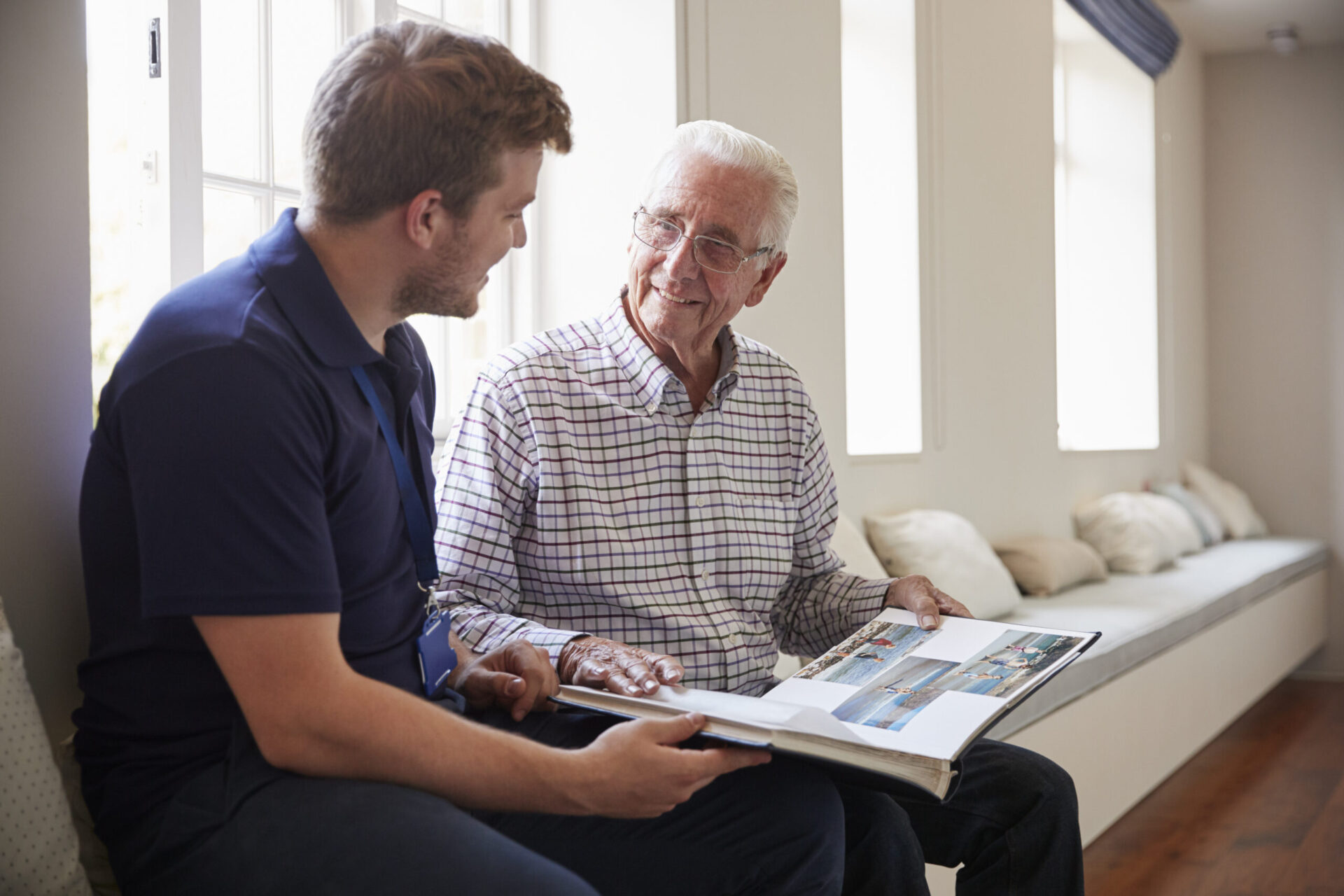 Senior man and caregiver looking at a photo album in a sunny room.
