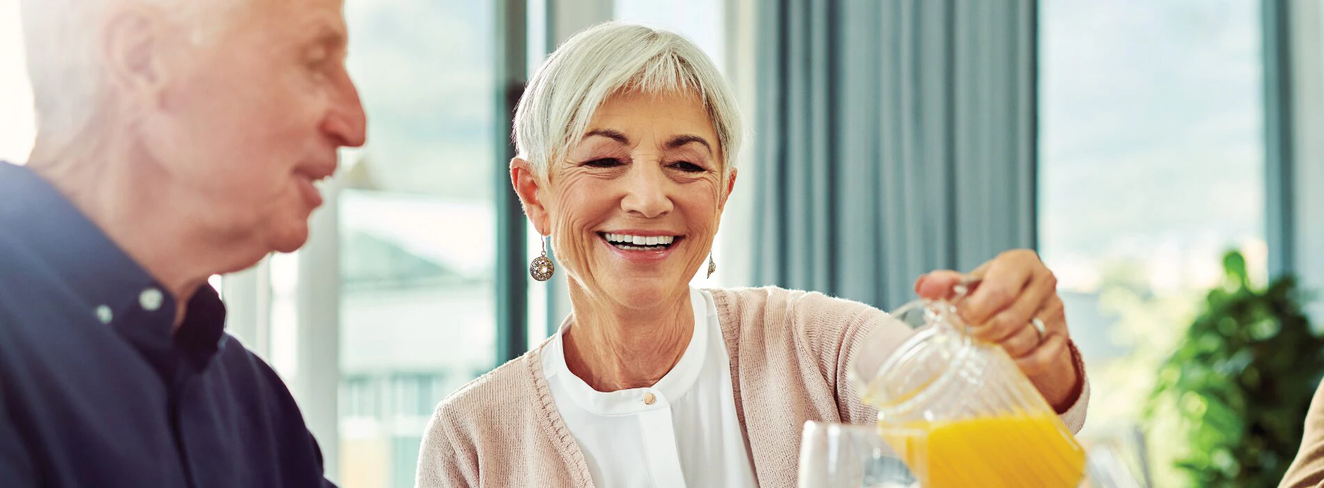 Elderly couple sitting and chatting at breakfast as senior woman pours a glass of orange juice.